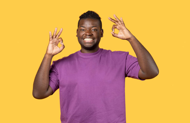 Portrait Of Positive Young Black Man Showing Ok Gesture At Camera Standing Over Yellow Studio Background, Gesturing Sign Of Approval With Both Hands. Everything Is Okay Concept - Photo, Image