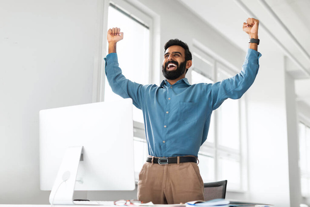 Jubelnder indischer Geschäftsmann, der am Arbeitsplatz im Büro vor dem Computer Erfolge feiert, Fäuste schüttelt und Ja schreit und emotional auf gute Nachrichten reagiert. Konzept zur großen Glücksfeier - Foto, Bild