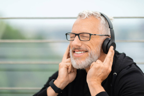 Schöne Musik zum Workout. Glücklicher älterer Mann mit Brille, der seine drahtlosen Kopfhörer berührt und mit geschlossenen Augen lächelt, während er im Freien trainiert, Kopierraum, Nahaufnahme - Foto, Bild
