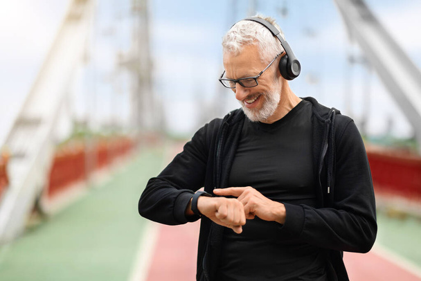 Happy retired sportsman exercising on footbridge, checking smart watch, copy space. Stopwatch, positive elderly man and runner check time, heart rate or monitor healthy body progress on data app - Φωτογραφία, εικόνα
