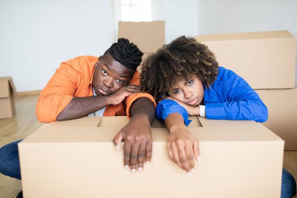 Расстроенные чернокожие супруги, склоняющиеся к картонной коробке, имеющие стресс при переезде в свой новый дом, истощенные афроамериканской пары страдают проблемы во время переселения, чувствуя усталость и депрессию - Фото, изображение