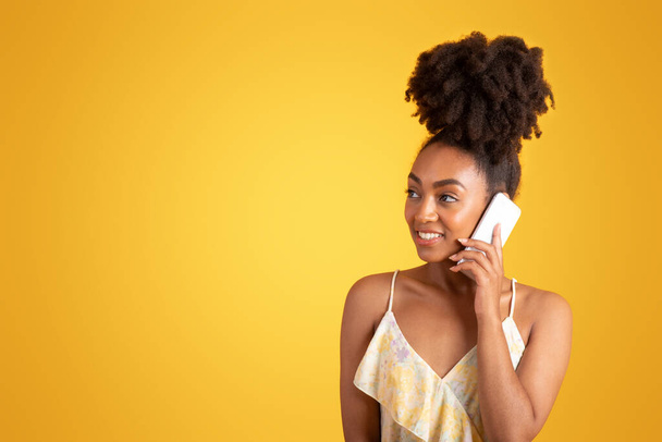 Happy millennial black woman calling by phone, thinking, looks at empty space, isolated on orange background, studio. Ad and offer, human emotions in spare time, gossip, good news and communication - Φωτογραφία, εικόνα