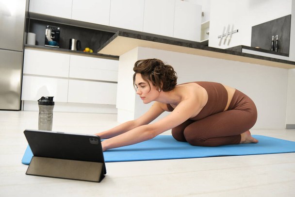 Kép a fiatal sportoló nő jógázik otthon, néz tornatanár bemutató videó tabletta, nyújtja test, fitness edzés gyakorlatok, pilates osztály online otthonról. - Fotó, kép