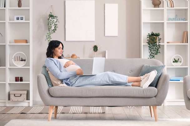 Молодая беременная женщина работает с ноутбуком на диване дома - Фото, изображение
