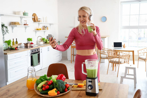 Спортивна зріла жінка з мобільним телефоном і склянкою здорової смуги на кухні - Фото, зображення