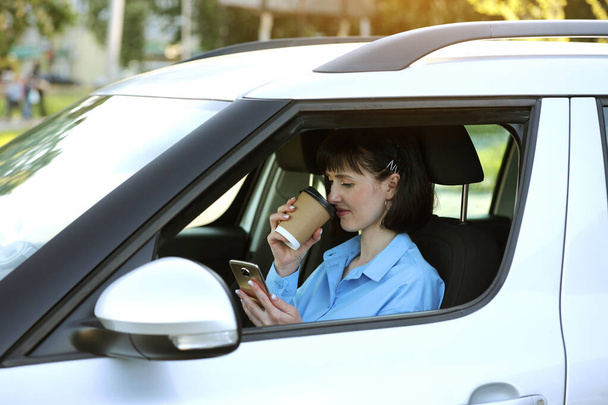 Uzaktan kumanda. Başarılı iş kadını girişimci, rahat bir arabada akıllı telefonla konuşurken elinde bir fincan kağıt kahve tutuyor. Trafik sıkışıklığının ortasında yolda çalışmak. Sonuç yönelimi kavramı. - Fotoğraf, Görsel