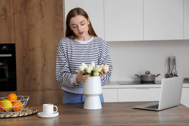 Hermosa mujer poniendo ramo de flores de tulipán blanco en jarrón en la mesa de madera en la cocina - Foto, imagen