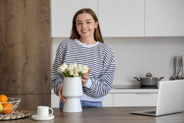 Feliz joven mujer sosteniendo jarrón con flores de tulipán blanco en la mesa en la cocina - Foto, Imagen