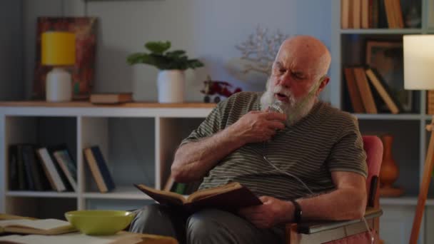 Vieil homme en masque respiratoire rester dans la chaise et lire le livre. Le vieil homme utilise un nébuliseur à cause de la respiration difficile. Thème de curation médicale de diverses maladies pulmonaires. Images 4k de haute qualité - Séquence, vidéo