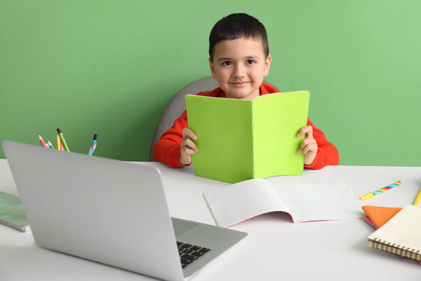 Netter kleiner Junge mit Textbuch macht Hausaufgaben am Tisch in der Nähe der grünen Wand - Foto, Bild