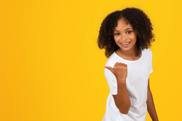Позитивна чорна дівчинка-підліток у білій футболці вказує пальцем на порожнє місце, рекомендує вивчити, ізольовано на жовтому тлі, студію. Схвалення продажу, школа, реклама та пропозиція, поради щодо освіти - Фото, зображення