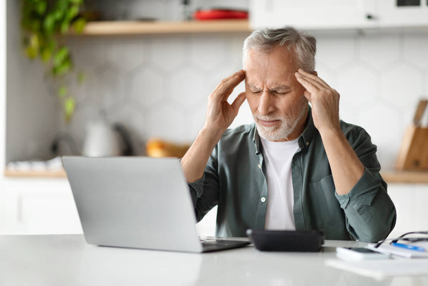 Porträt eines müden älteren Mannes, der an Kopfschmerzen leidet, während er zu Hause am Laptop arbeitet, ein kranker älterer Herr, der sich die Schläfen reibt und die Stirn runzelt, eine akute Migräne mit Bluthochdruck hat und am Schreibtisch in der Küche sitzt - Foto, Bild