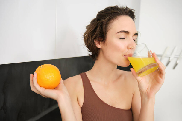 Estilo de vida saludable y deporte. Hermosa mujer sonriente, bebiendo jugo de naranja fresco y sosteniendo fruta en su mano, disfrutando de su bebida vitamínica después del entrenamiento en casa, de pie en la cocina. - Foto, imagen