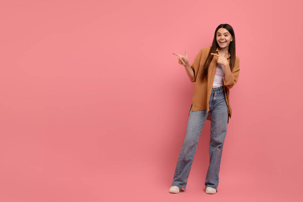 Хорошее предложение. Счастливая улыбающаяся девочка-подросток, указывающая на пространство для копирования двумя руками, веселая девушка-подросток, демонстрирующая свободное место для рекламы, стоя на розовом фоне, в полную длину - Фото, изображение