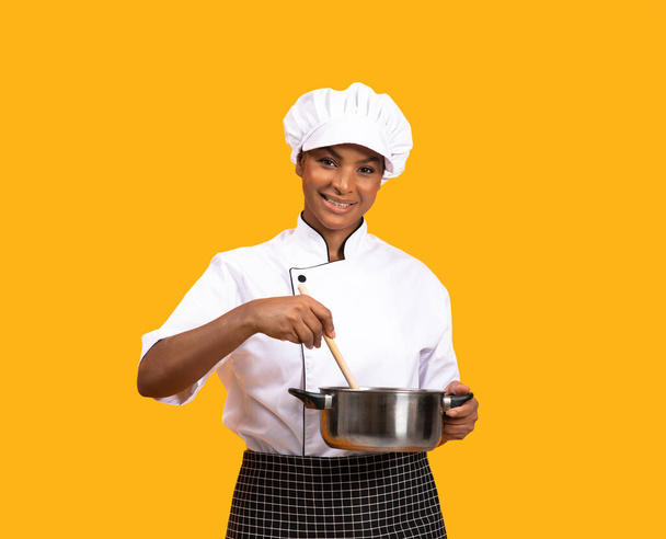 Красива чорна шеф-кухарка жінка з шпателем перемішуючи їжу в соусі і посміхаючись на камеру, щаслива молода афроамериканська кухарка леді стоячи ізольовані над жовтим фоном студії, копіювати простір - Фото, зображення