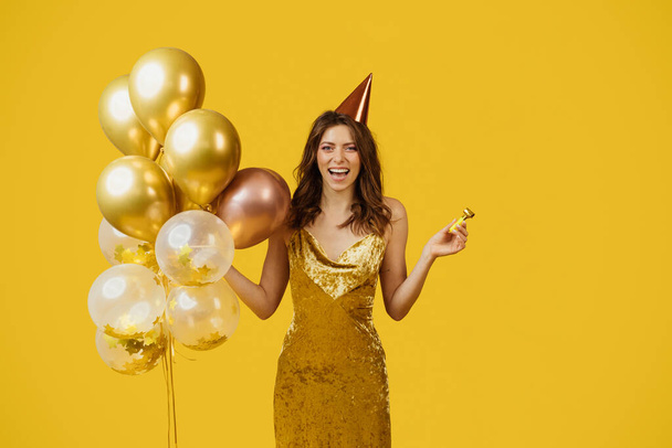 Tyytyväinen nainen mekko juhlava ilmapalloja, syntymäpäivälakki ja puolue puhallin juhlii erityinen tilaisuus, poseeraa yli keltainen studio tausta. Onnellinen nainen hauskaa lomalla - Valokuva, kuva