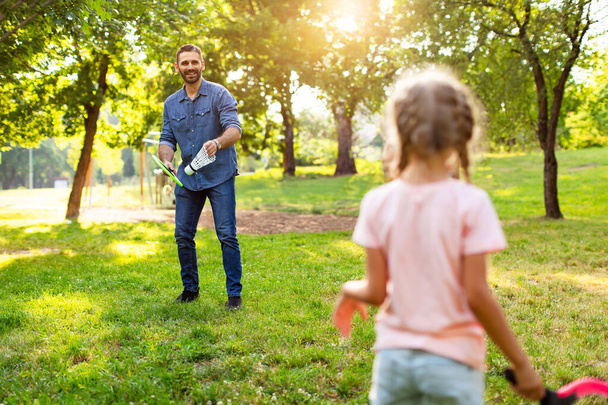 Rear view of middle aged man holding badminton racket and big shuttlecock while playing with his daughter outdoors in the park on summer day. Family pastime and values - Photo, Image