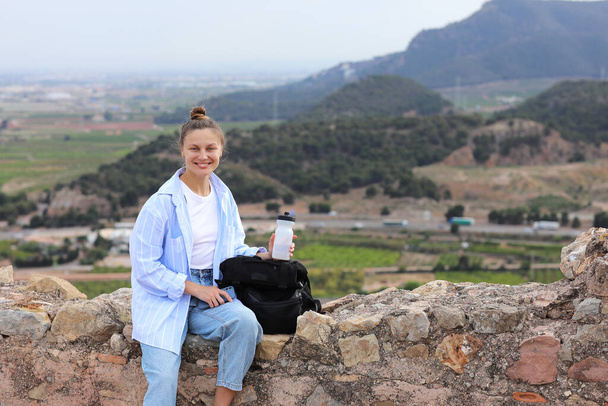 Gelukkige jonge vrouw reiziger met een rugzak houdt water fles zit op de heuvel tegen de achtergrond van een bergen na wandelen. Avontuur, zelfstandig reizen, toerisme, trekking, ecotoerisme op vakantie. - Foto, afbeelding