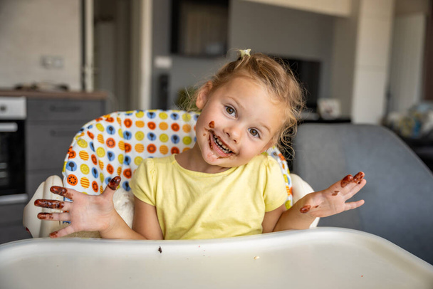 Little girl with blond hair eating homemade chocolate and showing mouth and dirty hands with stains of chocolate in home kitchen. High quality photo - Photo, Image