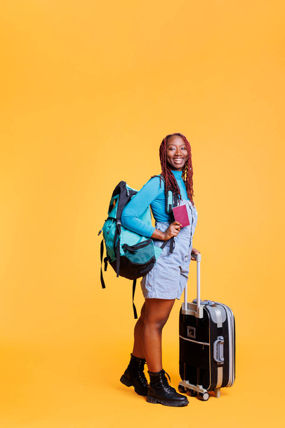 African american girl enjoying holiday trip with passport, carrying trolley bags luggage in studio. Young woman tourist travelling on urban adventure, weekend getaway with id documentation. - Photo, Image