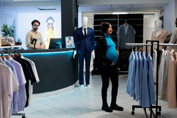 Jeune femme enceinte asiatique à la recherche de vêtements de maternité dans le centre commercial boutique de mode. Femme enceinte maman debout près de rack avec des vêtements suspendus dans le centre commercial, le choix du vêtement - Photo, image