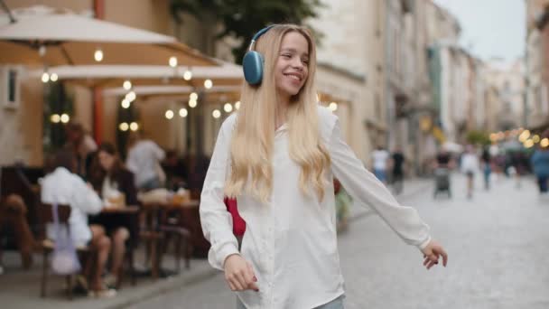 Happy overjoyed blonde young woman in wireless headphones listening favorite energetic music, celebrating victory win. Teenager girl tourist traveler walking in city street. Town lifestyles outdoors - Footage, Video