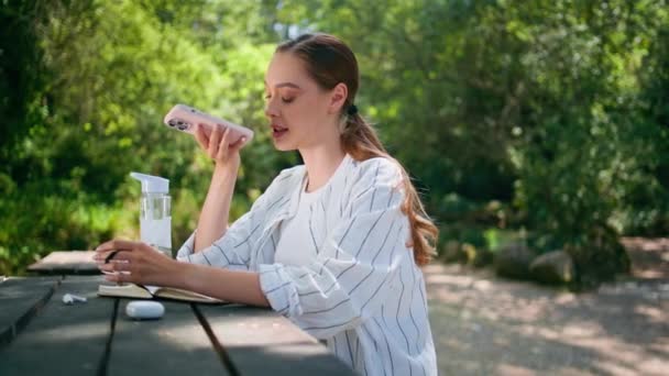 Girl recording audio message sitting beautiful forest table closeup. Relaxed young woman freelancer talking in smartphone loudspeaker at sunny nature. Attractive happy lady using voice recognition app - Footage, Video