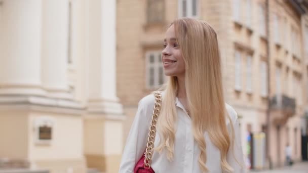 Portret młodej blondynki turystki spacerującej w miejskim tle ulicy w letni dzień. Nastolatka dziewczyna podróżnik uśmiechnięty o pozytywny nastrój korzystających na świeżym powietrzu. Miejski styl życia, wakacje - Materiał filmowy, wideo