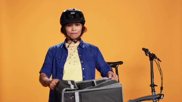 Kurye kadın müşteriye pizza getiren turuncu stüdyo arka planında izole edilmiş. BIPOC bisiklet sürücüsü müşteriye sipariş veriyor, termal sırt çantasından fast food kutuları çıkarıyor - Video, Çekim