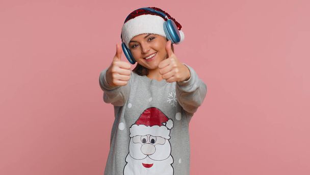 Ευτυχισμένη νεαρή γυναίκα των Χριστουγέννων στα ακουστικά ακούγοντας μουσική και χορεύοντας ντίσκο χαζεύοντας έχοντας διασκέδαση εκφραστική χειρονομίες χέρια, χαλαρώνοντας στο πάρτι. Υπέροχο κορίτσι απομονωμένο σε στούντιο ροζ φόντο - Φωτογραφία, εικόνα