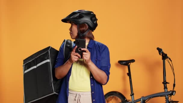 Confused asian woman looking around for directions, unsure of client location whereabouts. Professional courier wearing thermic backpack riding bike to deliver food, isolated over studio background - Footage, Video