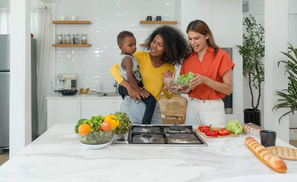 Two young diverse lesbian women spending time in a kitchen at home, harmoniously sharing food preparation duties whilst simultaneously nurturing a baby. - Photo, Image