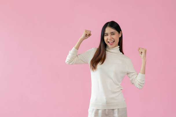 Um retrato de uma mulher asiática bonita e alegre em roupas casuais mostra seus punhos para cima e sorrindo para a câmera enquanto está de pé contra um fundo rosa isolado. - Foto, Imagem