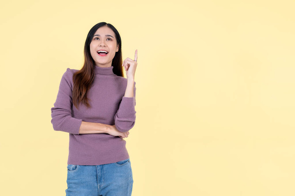 Een vrolijke en mooie Aziatische vrouw kijkt naar boven en wijst met haar vinger omhoog, staande tegen een geïsoleerde gele achtergrond. adviseren, adviseren, ideeën, opties, presentatie - Foto, afbeelding