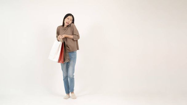 Egy gyönyörű, elfoglalt ázsiai nő beszél telefonon, miközben a bevásárlótáskáját cipeli. elszigetelt fehér háttér szöveges másolási hellyel. shopaholic, vásárlás életmód - Fotó, kép
