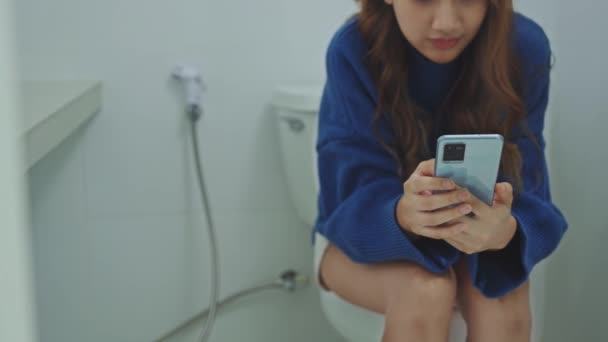 Mladá žena pomocí smartphone sedí na záchodové míse v domácí koupelně. - Záběry, video