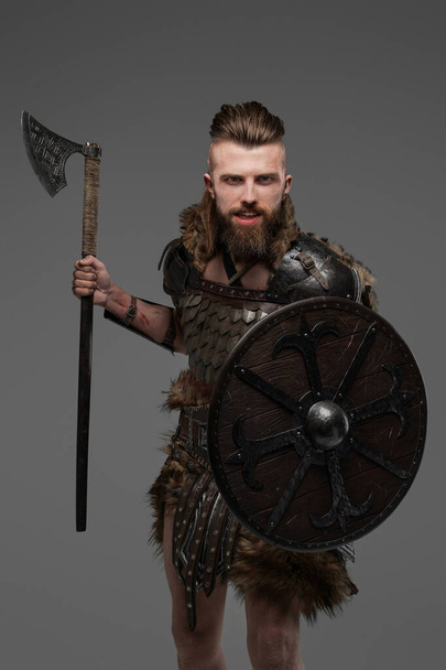 Fierce bearded Viking dressed in fur and light armor wielding an axe against a gray background - Photo, Image