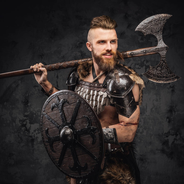 Um viking barbudo temível vestido com peles e armaduras leves, empunhando um machado e escudo contra uma parede cinzenta texturizada - Foto, Imagem