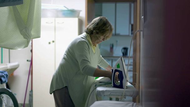 Una donna anziana stirare vestiti in piedi in lavanderia. Persona anziana candida che fa faccende domestiche quotidiane a casa. Autentica domesticità - Foto, immagini