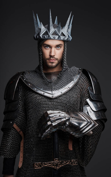 Портрет красивого короля в тяжелых доспехах, цепи и стальной короне, позирующего с мечом на сером фоне - Фото, изображение