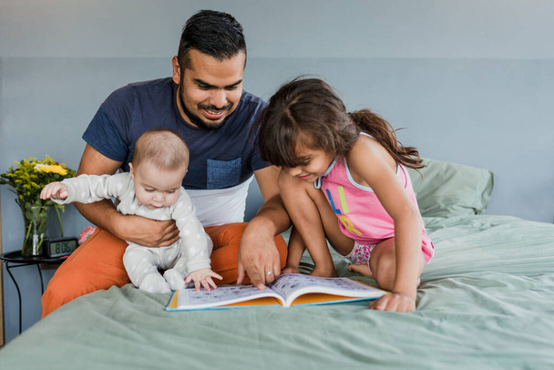 молодая латинская семья с детьми, читающих книгу вместе на кровати в Мексике, латиноамериканская мать, отец, дочь и ребенок в Латинской Америке - Фото, изображение