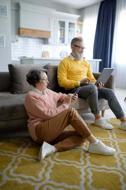 Glückliches Rentnerpaar mit digitalem Gerät, während es sich zu Hause im Wohnzimmer ausruht. Ältere Frau chattet mit Freunden auf Smartphone und älterer Mann kommuniziert in sozialen Medien - Foto, Bild