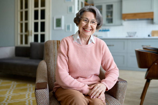 Портрет щасливої усміхненої літньої жінки, яка дивиться на камеру, сидячи на стільці у своїй домашній вітальні - Фото, зображення
