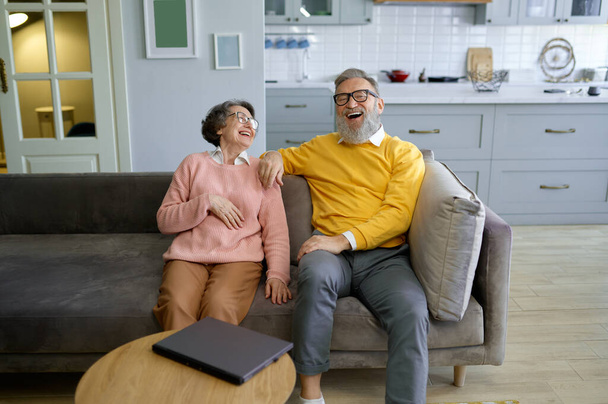 オンラインショッピングや予約旅行のためにラップトップを使用しながら,お互いを見つめ合うシニアカップルを笑う. 高齢者コンセプトによる現代技術 - 写真・画像