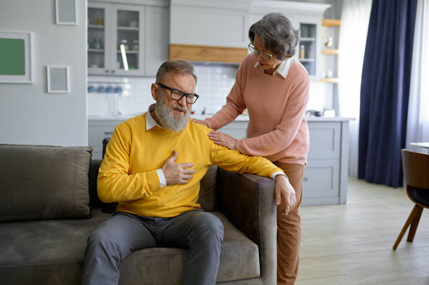 Starszy mężczyzna dotykający klatki piersiowej ma atak serca, a jego żona go wspiera. Koncepcja rodziny i opieki zdrowotnej w średnim wieku - Zdjęcie, obraz