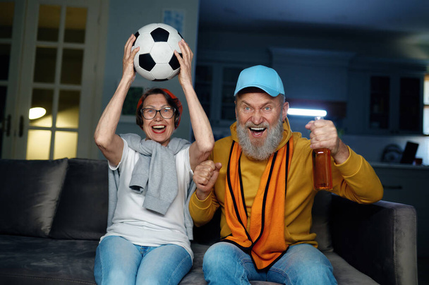 自宅でテレビでサッカーの試合を見ている楽しいシニアファミリーカップル. 高齢の夫と妻は,大喜びで興奮した叫び声を上げ,お気に入りのサッカーゲームを応援しています - 写真・画像