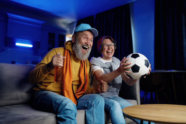 Expressieve senior familie koppel kijken voetbalwedstrijd op tv thuis. Ouderen man en vrouw gevoel dolblij en opgewonden schreeuwen en juichen favoriete voetbalwedstrijd - Foto, afbeelding