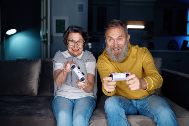 Senior couple playing video games together at home. Elderly wife and husband sitting on sofa holding joysticks and feeling excited of free time on weekend - Photo, Image