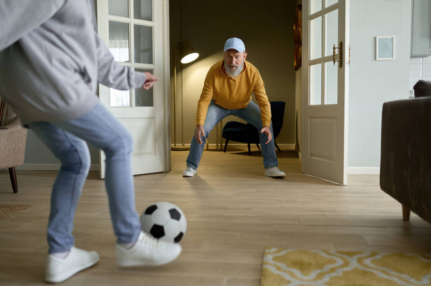 Счастливая пара на пенсии, играющая в футбол в гостиной. Пожилой муж пытается поймать мяч, пока старшая жена начинает пинаться. Развлечения в выходные - Фото, изображение