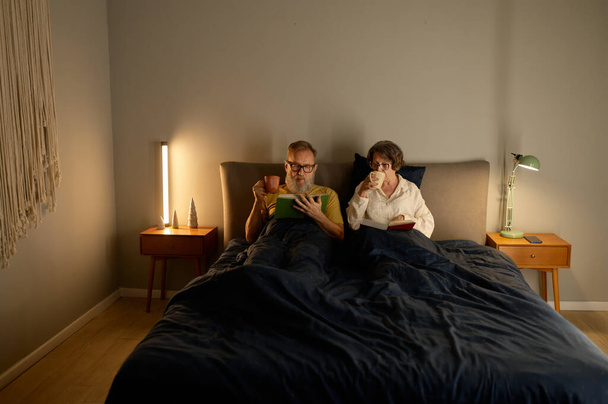 Calm family couple on retirement reading book together and drinking coffee while lying in bed before sleeping - Zdjęcie, obraz